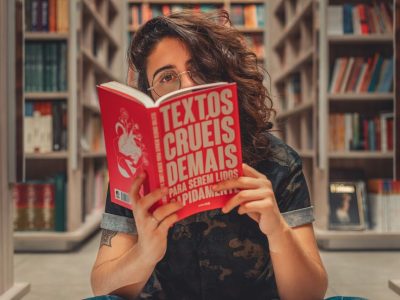 Jovem lê um livro sentada em uma biblioteca, cercada por estantes de livros.