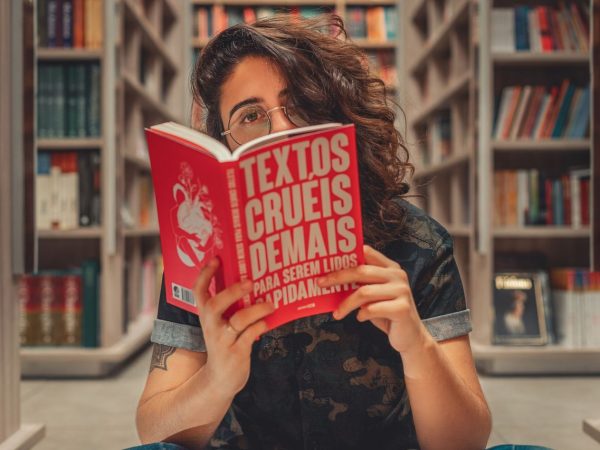Jovem lê um livro sentada em uma biblioteca, cercada por estantes de livros.