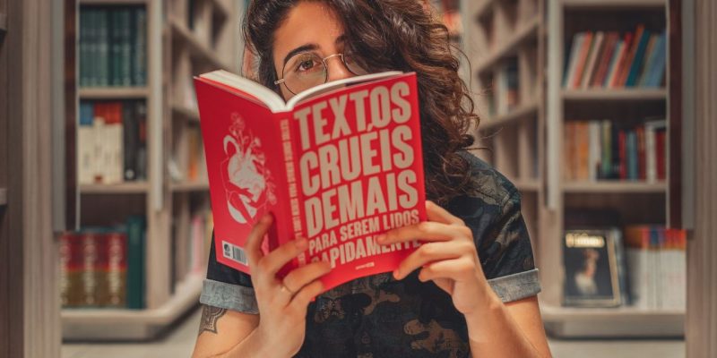 Jovem lê um livro sentada em uma biblioteca, cercada por estantes de livros.