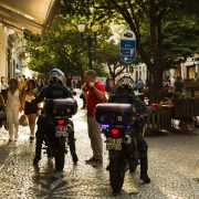 Agentes da guarda municipal em motocicletas patrulham uma rua movimentada, com pedestres ao redor.