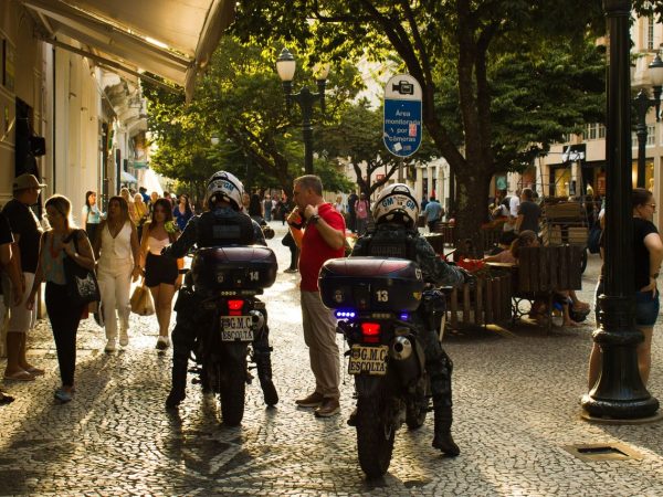 Agentes da guarda municipal em motocicletas patrulham uma rua movimentada, com pedestres ao redor.