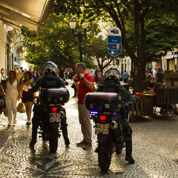 Agentes da guarda municipal em motocicletas patrulham uma rua movimentada, com pedestres ao redor.