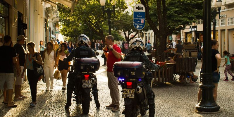 Agentes da guarda municipal em motocicletas patrulham uma rua movimentada, com pedestres ao redor.