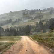 Estrada de terra atravessa uma paisagem montanhosa sob chuva e céu encoberto.