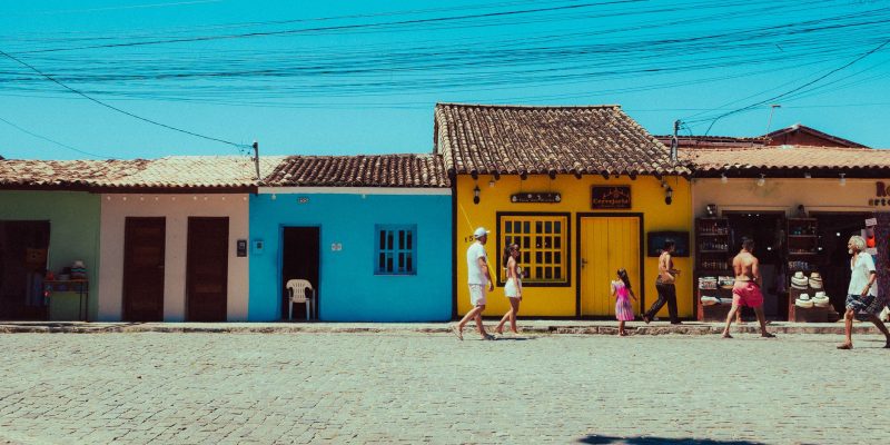 Pessoas caminham diante de construções tradicionais coloridas em um dia ensolarado.